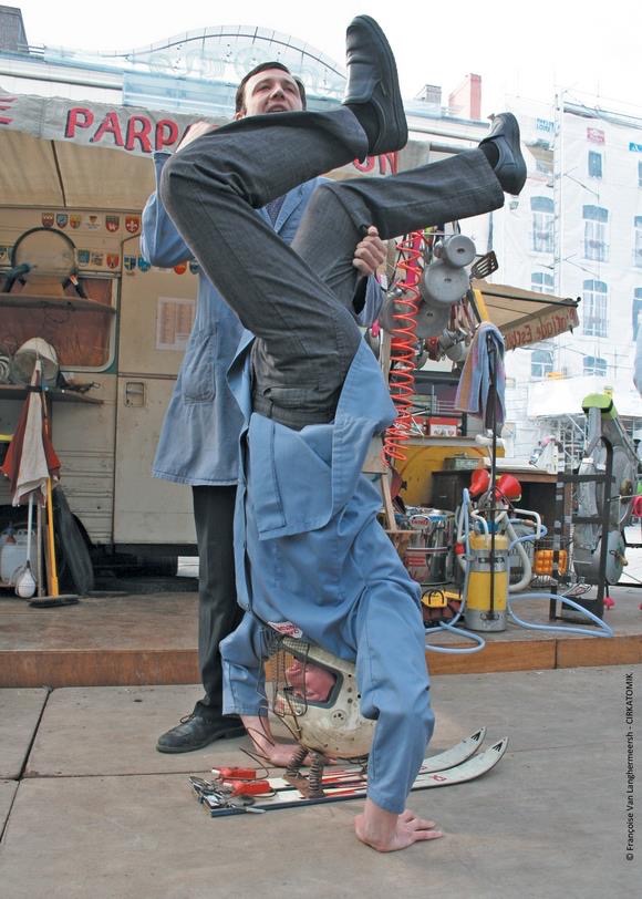 Deux comédiens en blouse bleue réalisent un numéro acrobatique en plein air, l'un en équilibre sur la tête, l'autre le soutenant.