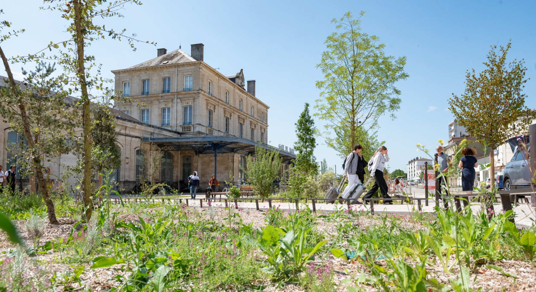 Esplanade réaménagée devant la gare de Niort avec jeunes arbres et plantations, des voyageurs avec bagages circulent