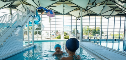 Un adulte accompagne un nourrisson dans le bassin intérieur d'un centre aquatique couvert, avec jouets gonflables colorés au-dessus de l'eau