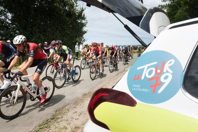 Peloton de cyclistes lancé sur une route de campagne, véhicule officiel Tour 79 au premier plan