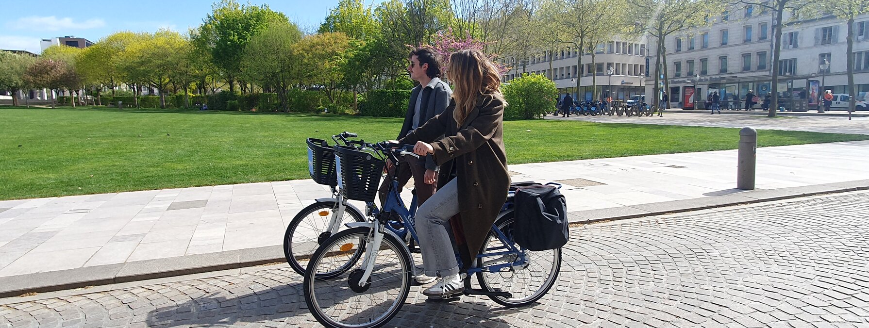Deux cyclistes, un homme et une femme, circulant côte à côte sur les pavés d'une place en plein soleil printanier