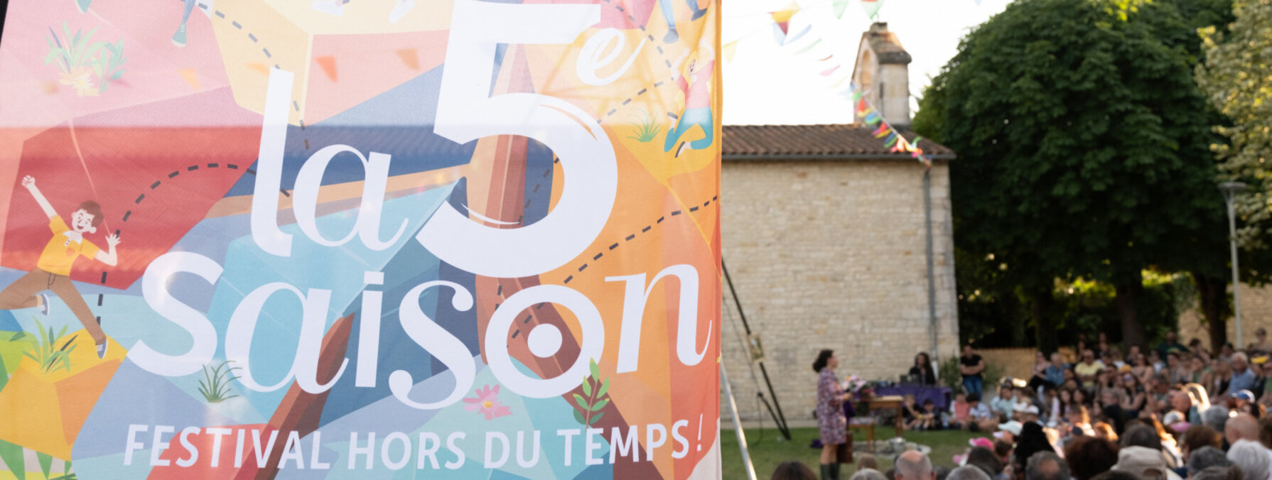 Affiche « Festival hors du temps » au premier plan, public nombreux assistant à un spectacle de plein air dans un cadre villageois