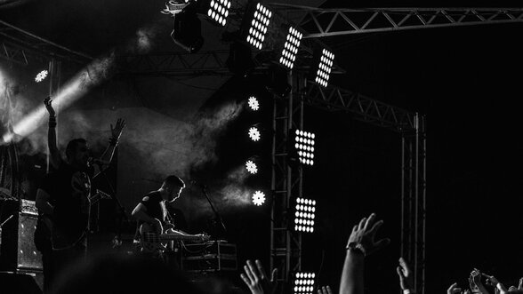 Artistes sur scène sous les projecteurs et la fumée, public les bras levés, ambiance de concert en noir et blanc