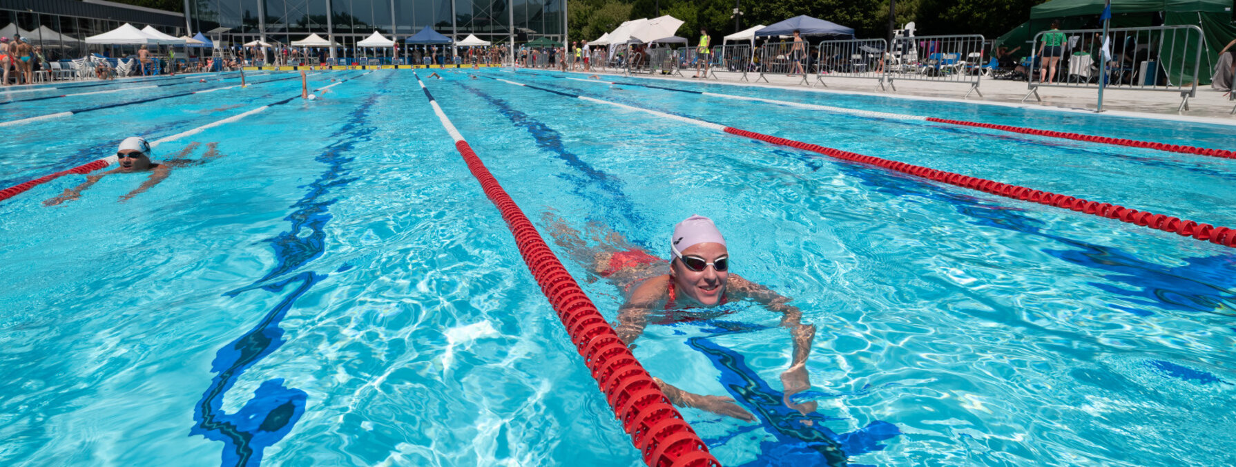 Nageurs dans les couloirs du bassin extérieur en plein soleil, avec le bâtiment de la piscine en fond
