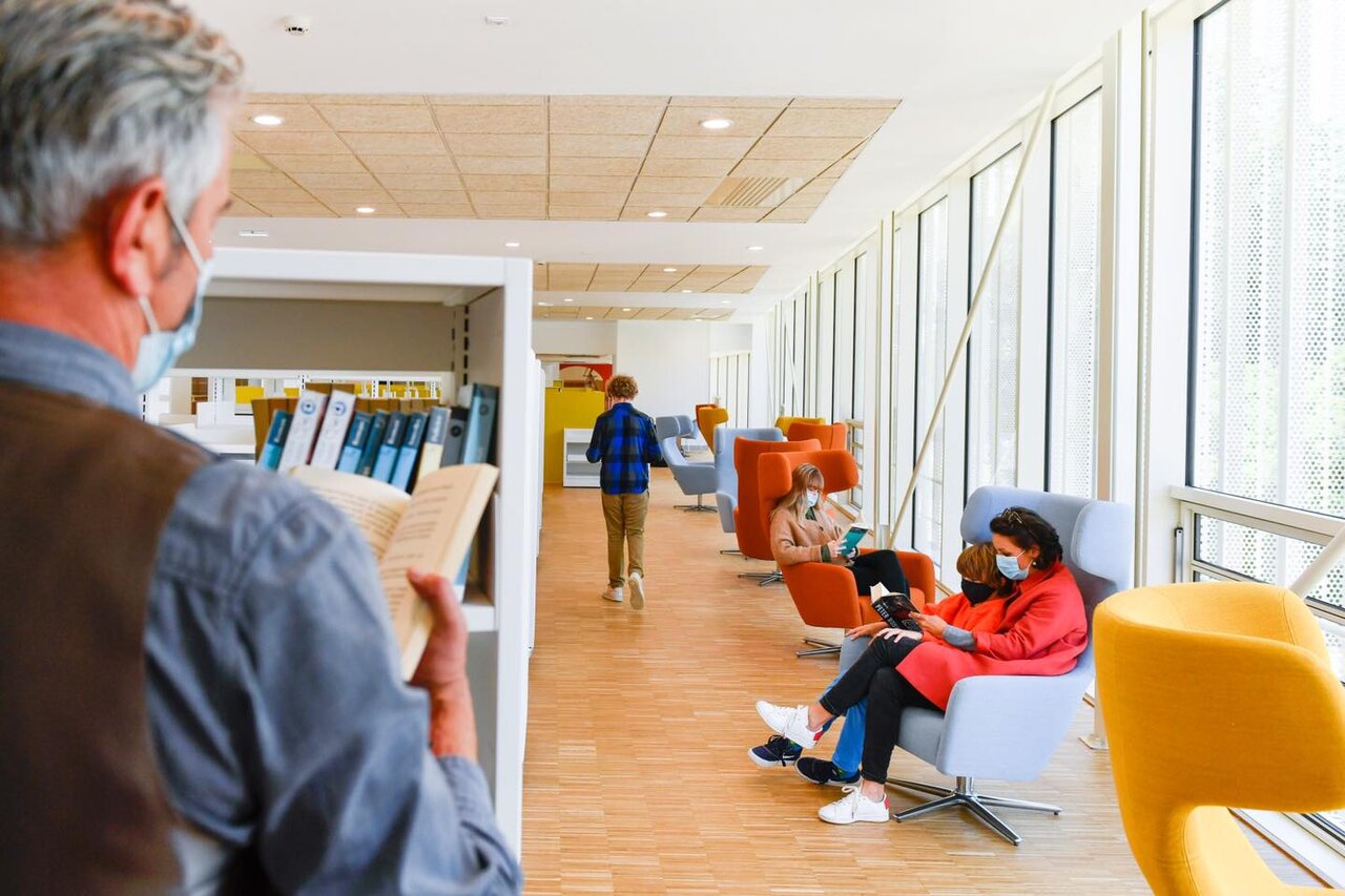 Usagers lisant en fauteuils colorés dans l'espace lumineux d'une médiathèque, un visiteur consultant des ouvrages en rayon