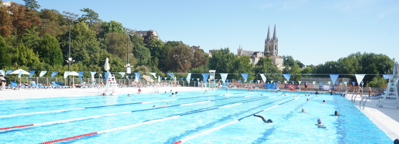Bassin extérieur de natation avec des nageurs dans les couloirs, l'église Saint-André de Niort visible en arriÚre-plan par temps ensoleillé