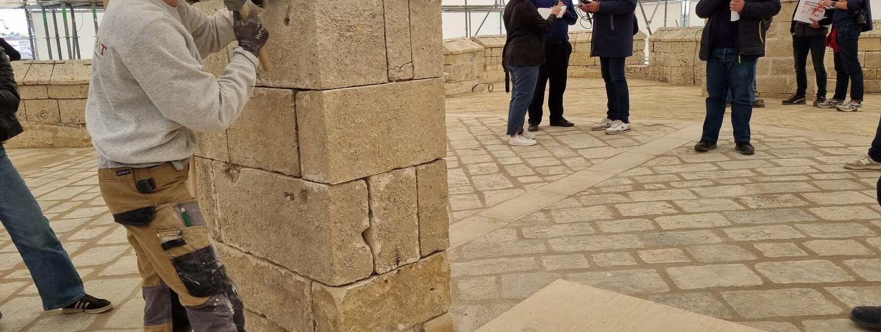 Une tailleuse de pierre façonne un pilier en tuffeau sous une tente de chantier, devant un groupe d'élus et journalistes casqués.