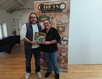 Deux personnes présentant la boîte de Circino 79 devant le kakémono du jeu « Le chasseur de trésors »