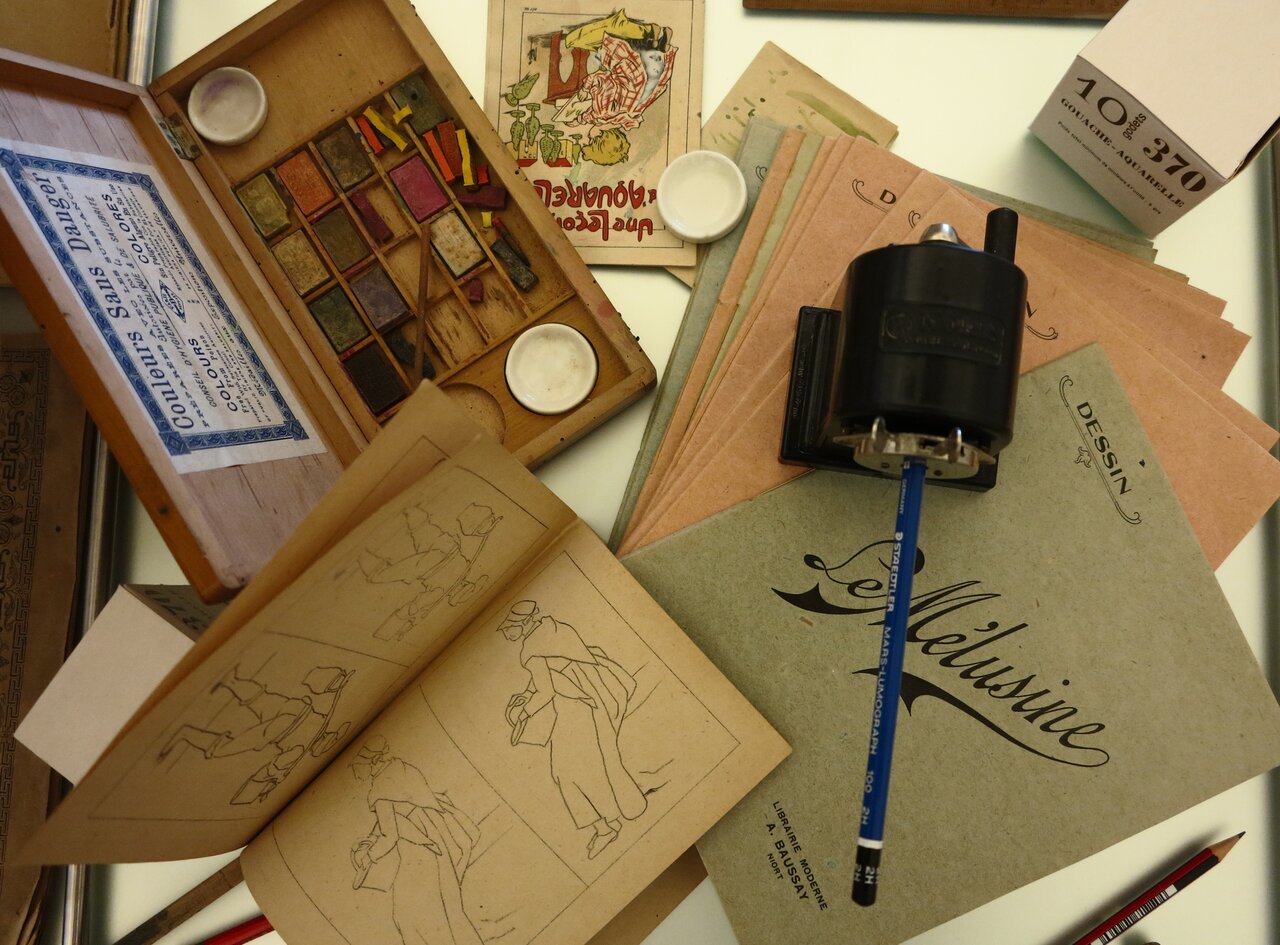 Boîte d'aquarelle ancienne, carnet de croquis ouvert sur des silhouettes et cahier « Le Mélusine » parmi des outils de dessin