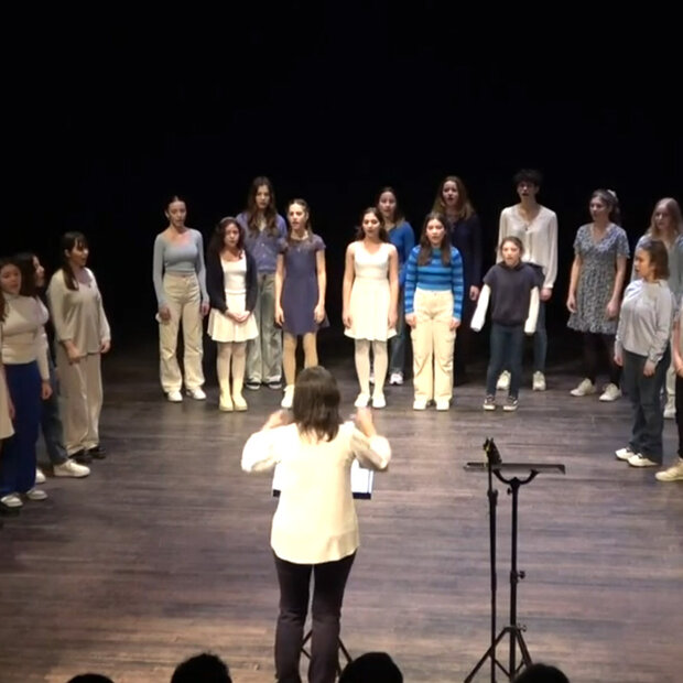 Des collégiens chantent en formation chorale sur scène, sous la direction d'une cheffe de chœur, accompagnés au piano