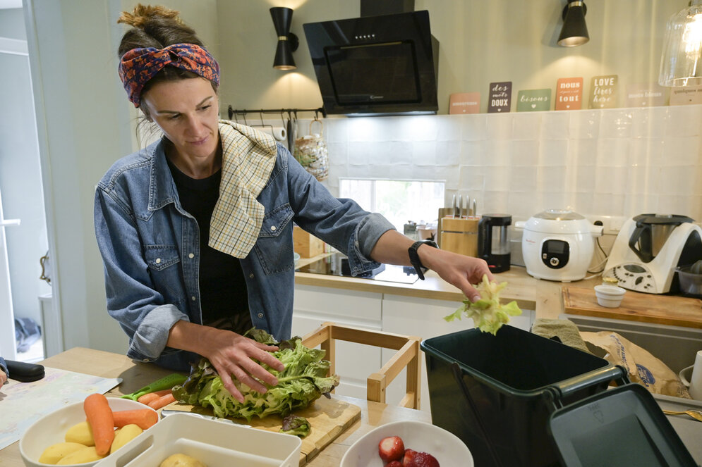Une personne dépose des feuilles de salade dans un bac de tri des biodéchets, entourée de légumes frais sur un plan de travail.