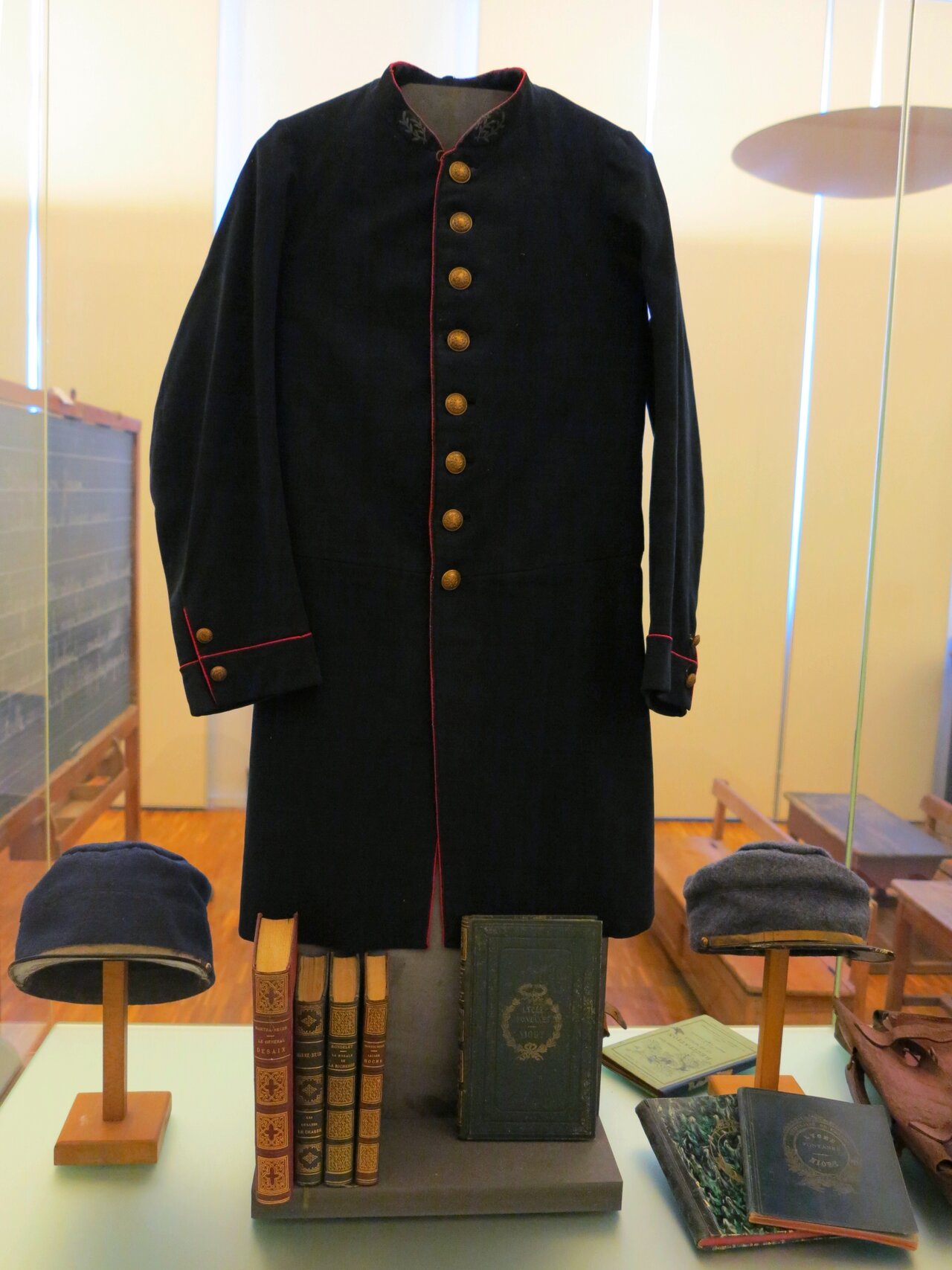 Vitrine de musée avec veste scolaire marine à boutons dorés et liseré rouge, deux casquettes sur présentoirs, livres et cartable en cuir