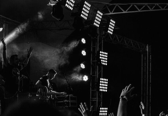 DJ sur scène en noir et blanc, entouré de lumières de concert et de mains levées dans le public