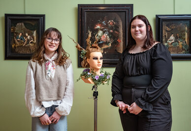 Deux apprenties coiffeuses posent dans une salle du musée encadrant une tête à coiffer ornée d'un chignon fleuri élaboré