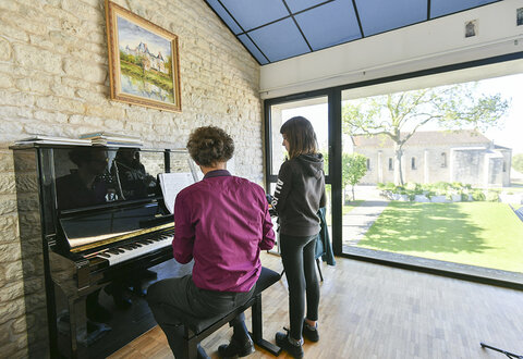 Professeur au piano accompagnant un élève lors d'un cours dans un studio lumineux donnant sur un jardin